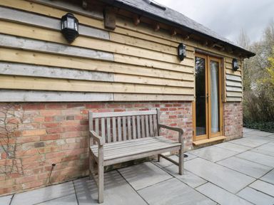 A wooden bench on a stone patio outside a brick and wooden building with glass double doors at The Courtyard - Hilltop Barn in Winterborne Zelston near Wareham
