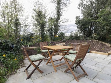 An outdoor patio with a wooden table and four wooden chairs with cushions surrounded by plants and trees at The Courtyard - Hilltop Barn in Winterborne Zelston near Wareham