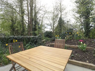 An outdoor wooden table with two wooden chairs in a garden with flowers and trees at The Courtyard - Hilltop Barn in Winterborne Zelston near Wareham