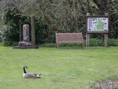 A park area with a wooden bench a stone marker a map stand and a Canada goose on the grass at The Courtyard - Hilltop Barn Winterborne Zelston near Wareham