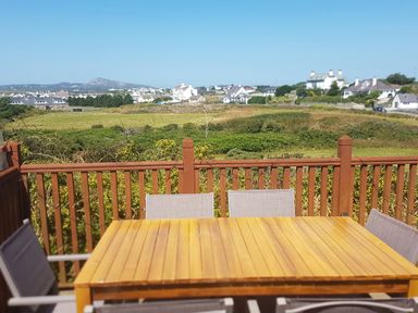 An outdoor wooden table with chairs on a balcony overlooking fields and houses at Windy Ridge in Trearddur Bay
