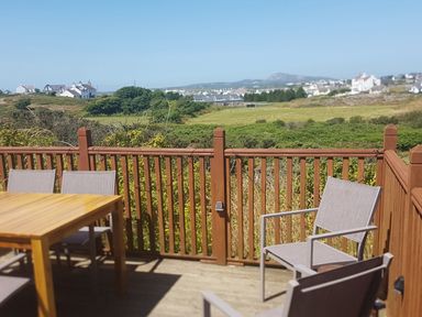 An outdoor wooden deck with table and chairs overlooking green fields and houses in the distance at Windy Ridge in Trearddur Bay