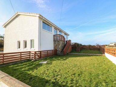 A fenced yard with grass and wooden stairs leading to a white two-story house at Windy Ridge in Trearddur Bay