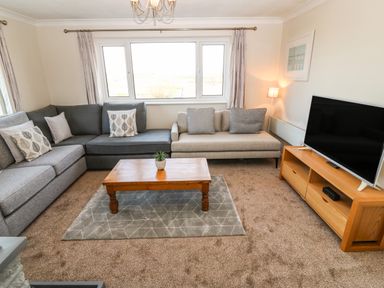 A living room with grey and beige sofas a wooden coffee table a grey rug a wooden TV stand and a TV at Windy Ridge in Trearddur Bay