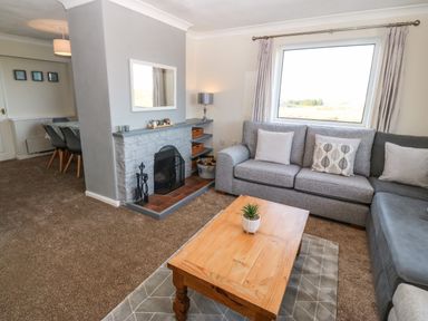 A living room with a gray sofa a wooden coffee table a stone fireplace and a dining area with gray chairs at Windy Ridge in Trearddur Bay