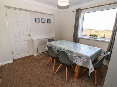 A dining room with a table covered by a patterned tablecloth and four chairs next to a window at Windy Ridge in Trearddur Bay