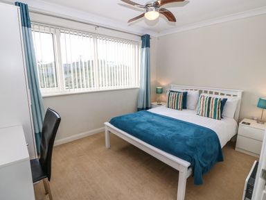 A bedroom with a double bed blue bedding and pillows two bedside tables with lamps a desk and chair and vertical blinds on the window at Windy Ridge in Trearddur Bay