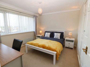A bedroom with a double bed yellow blanket two pillows two bedside tables with lamps a wooden desk with a black chair and a large window with vertical blinds at Windy Ridge in Trearddur Bay