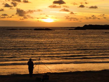 A person fishing at the shore during sunset with the sea and small islands in the background at Windy Ridge in Trearddur Bay