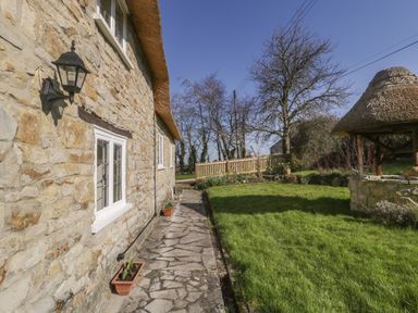 A stone house with a thatched roof beside a stone paved pathway and a grassy garden with a wooden fence and thatched structure at Field View in Curland near Taunton