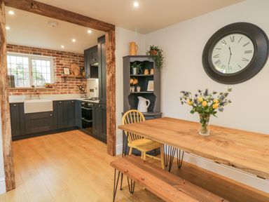 A kitchen with dark cabinets and brick wall next to a dining area with wooden table bench and chair at Pumphouse Cottage in Uffculme