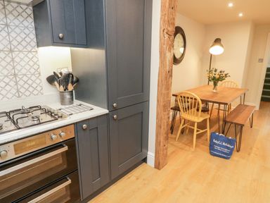 A kitchen area with a gas stove and dark cabinets next to a dining table with chairs and a bench at Pumphouse Cottage in Uffculme