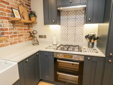 A kitchen corner with dark cabinets a built in oven and stove a brick wall with a wooden shelf and various kitchen items at Pumphouse Cottage in Uffculme