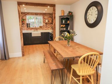 A dining area with a wooden table, yellow chairs, a kitchen with dark cabinets and a brick wall at Pumphouse Cottage in Uffculme