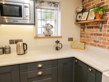 A kitchen corner with dark cabinets a microwave kettle coffee press and a window with cups on a rack at Pumphouse Cottage in Uffculme