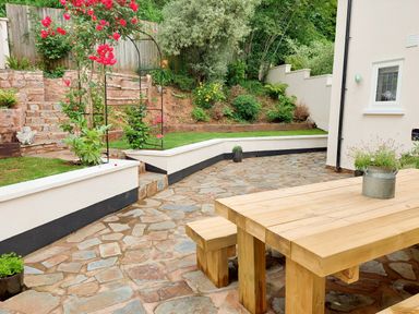 A garden patio with a wooden table and benches stone steps and planted flowers at Pumphouse Cottage in Uffculme
