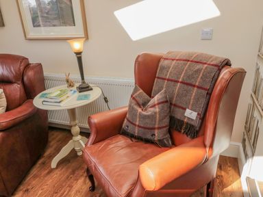 A leather armchair with a plaid throw and matching cushion next to a small round side table with books and a lamp at Cushat House in Stamford near Embleton