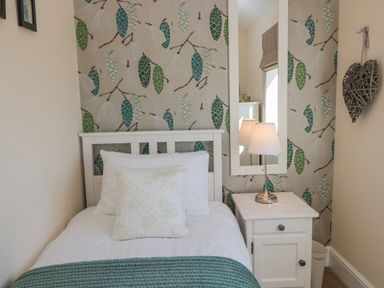 A bedroom with a single bed with white bedding and a green throw a white bedside table with a lamp a tall mirror and leaf patterned wallpaper at Cushat House Stamford near Embleton