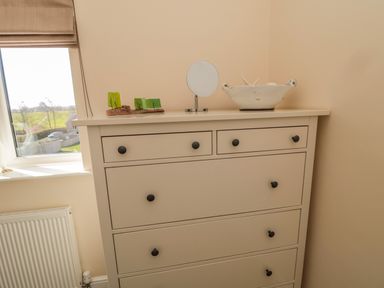 A beige chest of drawers with a small round mirror and decorative items on top near a window with a beige blind at Cushat House Stamford near Embleton