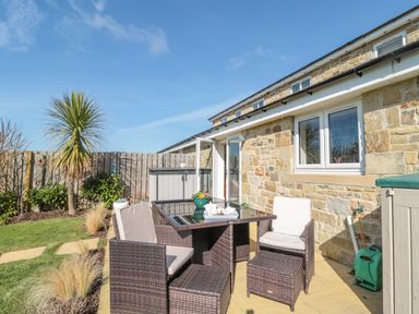 An outdoor patio with a glass table and four cushioned chairs next to a stone house with a garden and blue sky at Cushat House Stamford near Embleton