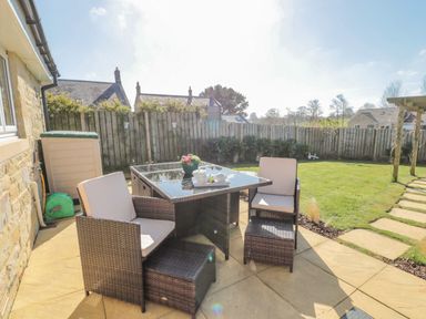 An outdoor patio with a glass top table and four wicker chairs with cushions in a fenced garden with grass and houses in the background at Cushat House Stamford near Embleton