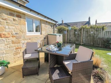 An outdoor patio with a glass table and four wicker chairs with cushions next to a stone wall and wooden fence at Cushat House Stamford near Embleton