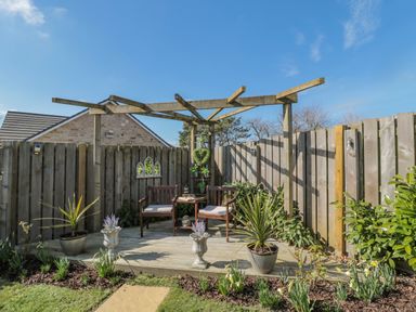 A wooden deck with two chairs and a small table under a pergola in a fenced garden with potted plants and flowers at Cushat House Stamford near Embleton