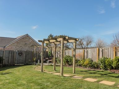 A garden with wooden pergola and stone pathway leading to seating area surrounded by plants and fence at Cushat House Stamford near Embleton