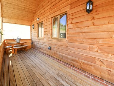 A wooden porch with a wooden table and bench seating with cushions and a watering can at The Larches in Van near Llanidloes