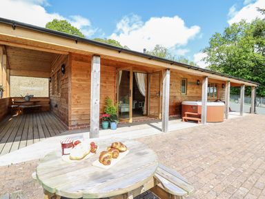 An outdoor wooden table with croissants bread and jam in front of a wooden building with open doors and a hot tub at The Larches Van near Llanidloes