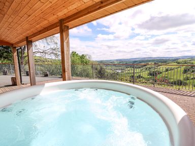 A hot tub under a wooden roof overlooking a valley with green hills and a river at The Larches Van near Llanidloes
