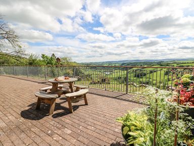 An outdoor patio with a wooden picnic table and benches overlooking a green valley with a metal railing at The Larches Van near Llanidloes