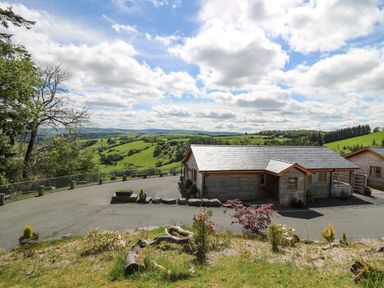 A wooden cabin with a paved driveway overlooking green hills and a partly cloudy sky at The Larches in Van near Llanidloes