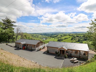 Two detached wooden buildings with slate roofs on a paved area overlooking green rolling hills at The Larches in Van near Llanidloes