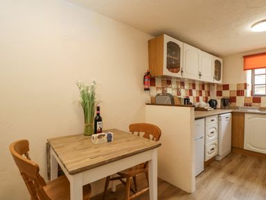 A small kitchen with a wooden table and chairs a bottle a plant and biscuits on the table and cabinets appliances and a window with an orange blind at The Old Bakery in South Perrott near Mosterton