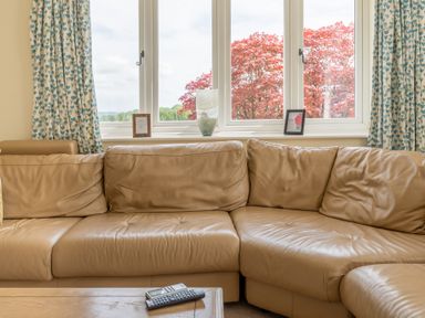 A beige leather sectional sofa in front of a window with blue patterned curtains and a wooden table with remote controls at Hazelseat House in Graythwaite near Ulverston
