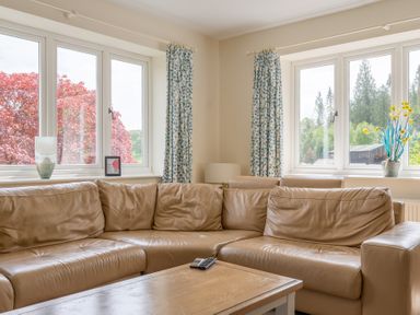 A living room with a tan leather corner sofa near windows with patterned curtains and a wooden coffee table at Hazelseat House in Graythwaite near Ulverston