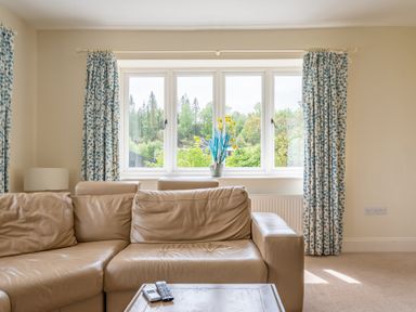 A beige leather sofa with a wooden table holding remote controls in front of a window with patterned blue and white curtains and a vase of yellow flowers at Hazelseat House in Graythwaite near Ulverston