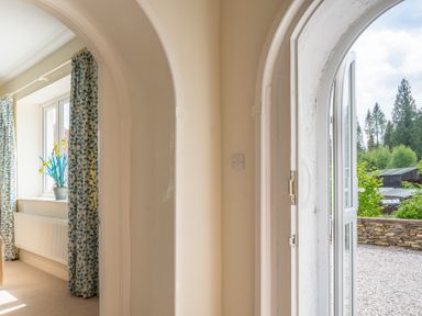 Arched hallway with a window having flower curtains and a potted plant and an open arched door showing an outdoor gravel area and trees at Hazelseat House in Graythwaite near Ulverston