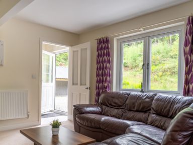 A living room with a brown leather sofa purple curtains wooden coffee table and an open door showing the outside at Hazelseat House in Graythwaite near Ulverston