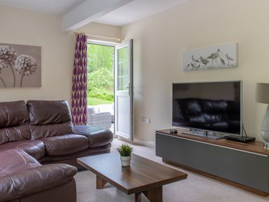 A living room with a brown leather sofa a wooden coffee table a flat screen tv on a cabinet and a door open to a garden at Hazelseat House in Graythwaite near Ulverston