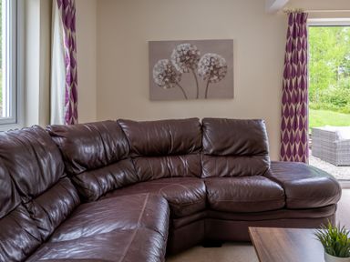 A room with a brown leather sectional sofa purple and white curtains and a painting of flowers at Hazelseat House Graythwaite near Ulverston