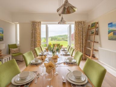 A dining room with a wooden table set for eight people with green chairs and view of landscape at Hazelseat House in Graythwaite near Ulverston
