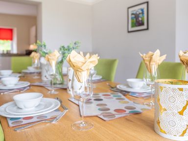 A dining table set with white plates bowls silverware glasses with napkins and placemats in a room with green chairs and wall art at Hazelseat House in Graythwaite near Ulverston