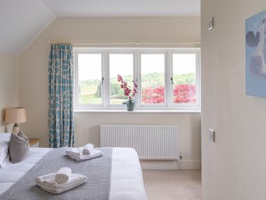 A bedroom with a bed showing rolled towels on a gray knitted throw beside a bedside table with a lamp and a window with framed curtains and a potted flower at Hazelseat House in Graythwaite near Ulverston