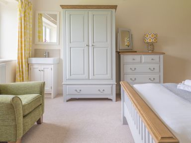 A bedroom with a green armchair a white wardrobe a white chest of drawers with a lamp and a small mirror a bed with folded towels and a sink with yellow patterned curtains at Hazelseat House in Graythwaite near Ulverston