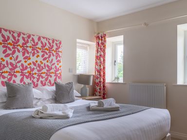 A bedroom with a bed with grey pillows and towels a bedside table with lamp and windows with curtains at Hazelseat House in Graythwaite near Ulverston