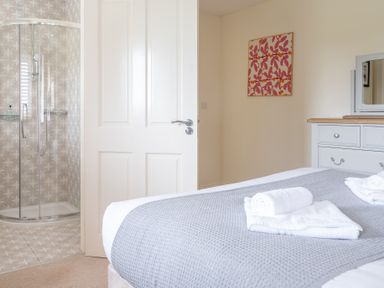 A bedroom with a bed covered with a gray blanket and white towels with a dresser and an open door showing a shower at Hazelseat House Graythwaite near Ulverston