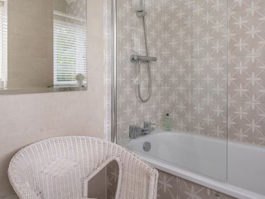 A bathroom with a bathtub a showerhead a glass shower screen a wicker chair and a mirror at Hazelseat House Graythwaite near Ulverston