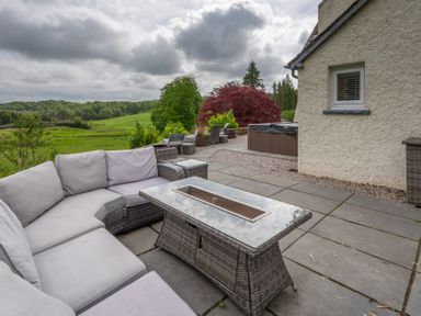 An outdoor patio with a wicker sectional sofa and table near a beige house with a view of trees and green fields at Hazelseat House in Graythwaite near Ulverston